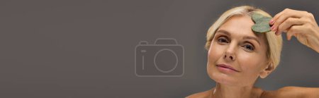Foto de Preciosa mujer madura usando gua sha sobre un fondo gris. - Imagen libre de derechos
