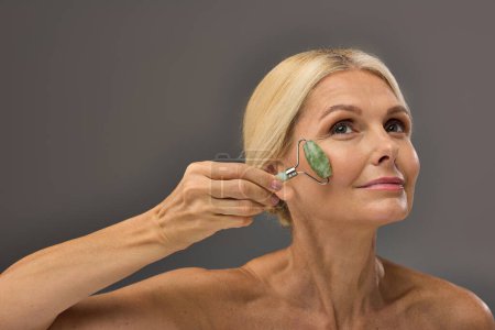 Gorgeous mature woman using face roller on a gray backdrop.