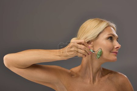 Pretty mature woman using face roller on a gray backdrop.
