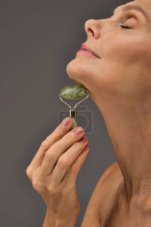 Attractive mature woman using face roller on a gray backdrop.