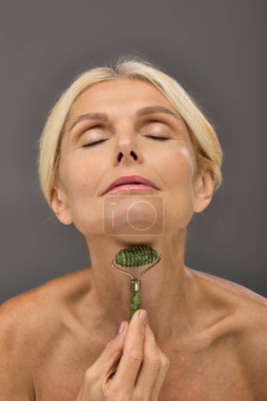 Foto de Lovely mature woman using face roller on a gray backdrop. - Imagen libre de derechos