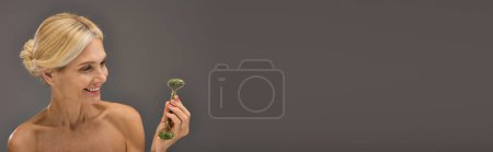 Téléchargez les photos : Belle femme mature à la recherche en utilisant un rouleau de visage sur une toile de fond grise. - en image libre de droit