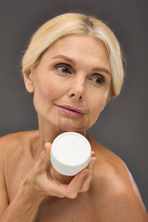 Mature woman gracefully holds a jar of cream in her hand on a gray backdrop.
