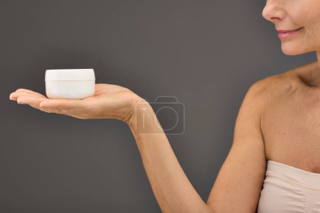Téléchargez les photos : Femme mûre tenant élégamment une tasse de crème pour la routine des soins de la peau. - en image libre de droit