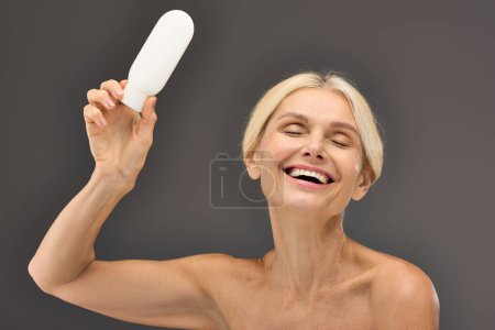 Téléchargez les photos : Charming mature woman holding locion on a gray backdrop. - en image libre de droit