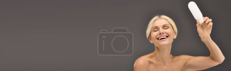Foto de Mujer madura alegre sosteniendo loción sobre un fondo gris. - Imagen libre de derechos