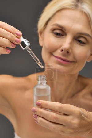 Photo for Charming mature woman applying serum on a gray backdrop. - Royalty Free Image