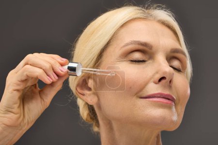 Téléchargez les photos : Gorgeous mature woman applying serum on a gray backdrop. - en image libre de droit