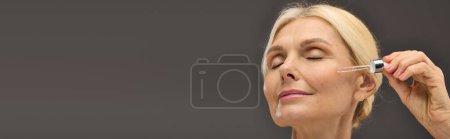Preciosa mujer madura aplicando suero sobre un fondo gris.