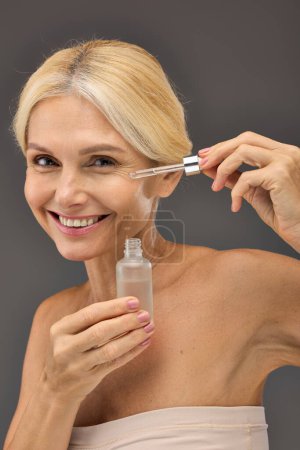 Photo for Good looking mature woman applying serum on a gray backdrop. - Royalty Free Image