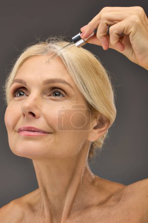 Photo for Jolly mature woman applying serum on a gray backdrop. - Royalty Free Image
