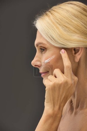 Téléchargez les photos : Femme appliquant gracieusement de la crème sur son visage sur un fond gris. - en image libre de droit
