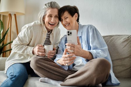 Téléchargez les photos : Deux femmes âgées assises sur un canapé, prenant joyeusement un selfie ensemble. - en image libre de droit