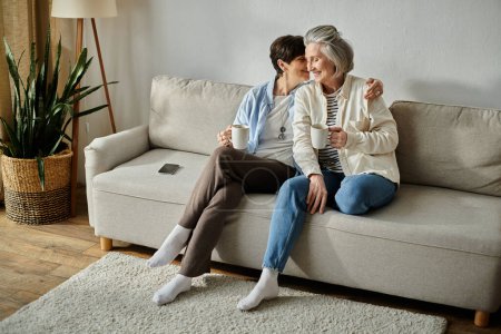 Zwei ältere Menschen, ein reifes lesbisches Liebespaar, sitzen zusammen auf einer Couch und genießen Tassen Kaffee.