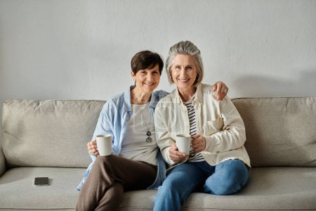 Two senior women relax on a couch, sipping coffee from mugs.