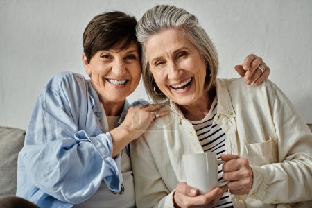 Two older women, a loving mature lesbian couple, sit together on a couch, enjoying a cup of coffee.
