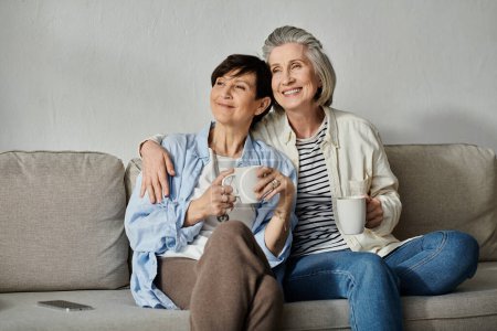 Two older women enjoying coffee together on a cozy couch.