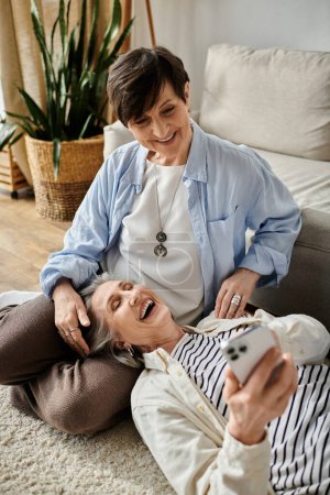 Foto de Dos mujeres se ríen sentadas en el suelo, absortas en un teléfono celular. - Imagen libre de derechos