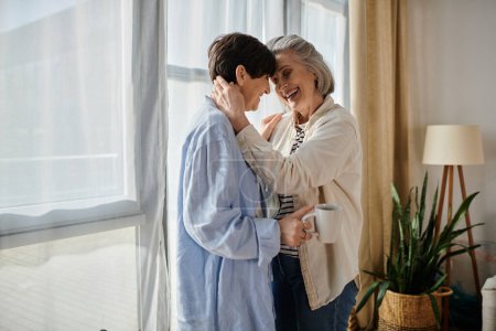 Two elderly women share a heartwarming hug in front of a window.