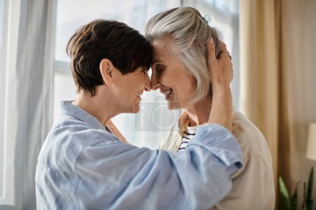 Two older women embrace lovingly in front of a window.