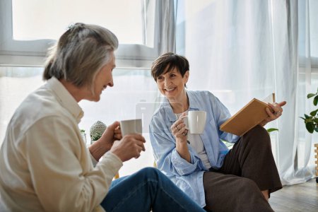 Téléchargez les photos : Two elderly individuals, a loving mature lesbian couple, sit on the floor, sipping coffee. - en image libre de droit