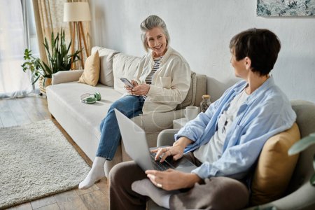 Photo for Two people, a loving mature lesbian couple, sit on a couch and use a laptop. - Royalty Free Image