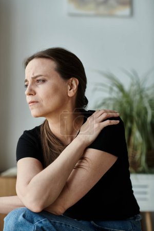 A middle-aged woman seated, arm on shoulder, deep in thought.
