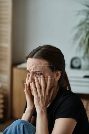 A middle-aged woman sits at home, covering her face with her hands in distress.