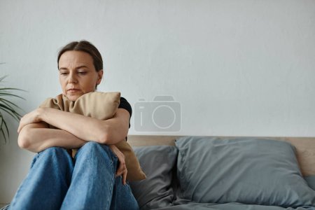 Téléchargez les photos : A middle-aged woman sits on a bed, arms crossed, showing resilience. - en image libre de droit