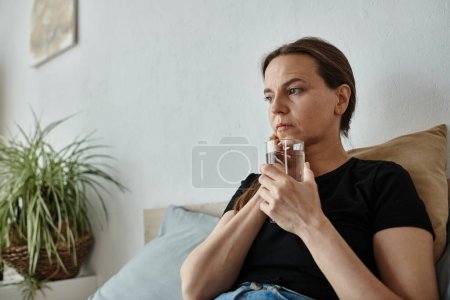 Photo for A middle-aged woman sits on a bed holding a glass of water, lost in thought. - Royalty Free Image