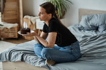 Middle-aged woman sits on bed with birthday cake.