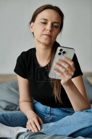 Middle-aged woman sitting on bed, absorbed in smartphone.