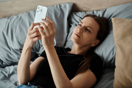 A middle-aged woman lying in bed, engrossed in her phone screen.
