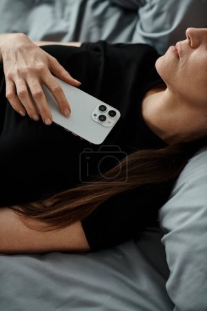 Middle-aged woman in bed with phone, seeking solace and support through online therapy during a mental breakdown.
