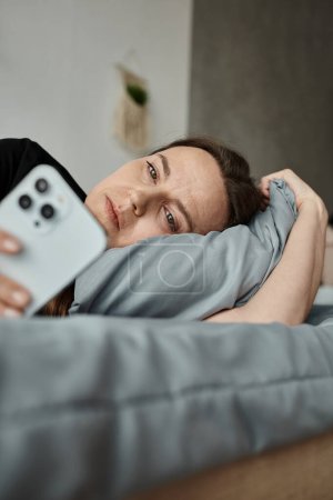 A woman resting in bed, engrossed in her phone screen.
