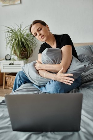 Middle-aged woman sits on bed, engrossed in laptop.