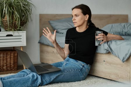 Middle-aged woman sits with laptop and water, absorbed in online therapy session.