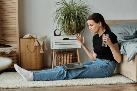 Middle aged woman sitting on floor, engaging in online therapy with laptop and staying hydrated with water bottle.