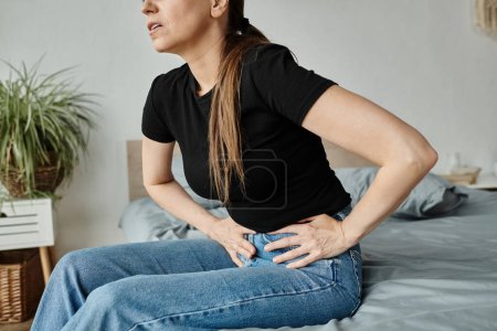 Middle-aged woman sitting on bed grimacing in pain, clutches stomach.