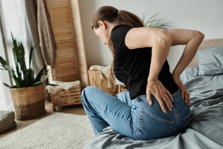 Téléchargez les photos : A middle-aged woman sits on a bed, experiencing back pain, struggling with depression. - en image libre de droit
