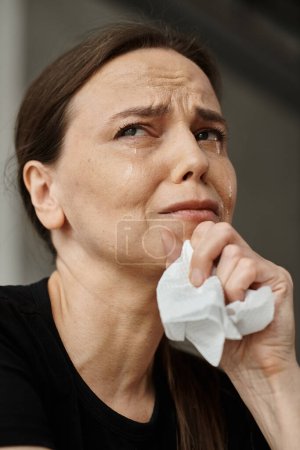 Middle-aged woman wipes her face with a tissue, dealing with mental distress.