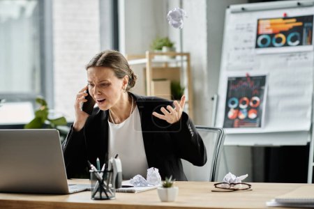 Middle-aged woman talking on phone at office desk, overwhelmed by stress.