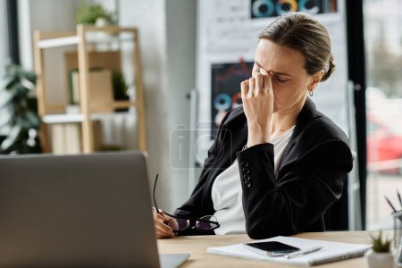 Foto de A middle-aged woman sits at a desk with a laptop, showing signs of stress and mental strain. - Imagen libre de derechos