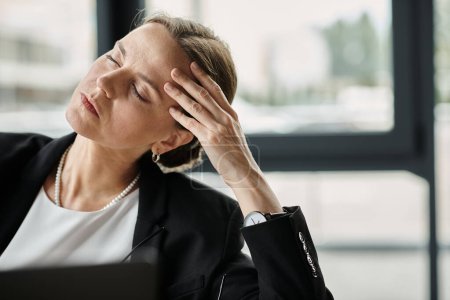 Middle aged woman in business suit holds head while working on laptop.