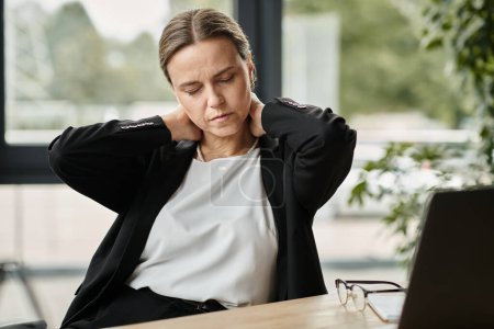 Téléchargez les photos : Middle-aged woman with neck pain, stressed, sitting in front of laptop. - en image libre de droit