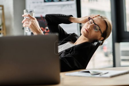 Woman holds water near laptop in stressful office environment.