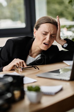 Stressed middle-aged woman rests head on desk in office.