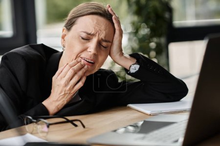 Stressed middle-aged woman sits at her desk with hand on head.