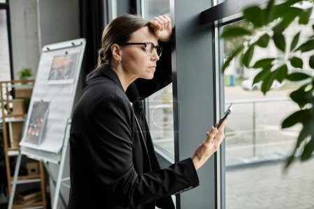 Middle-aged woman in glasses leans against window, looking at phone screen.