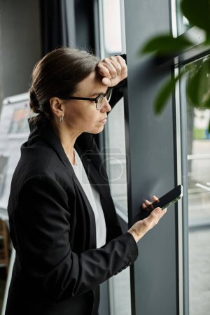 A middle-aged business woman stands by a window, absorbed in her phone.